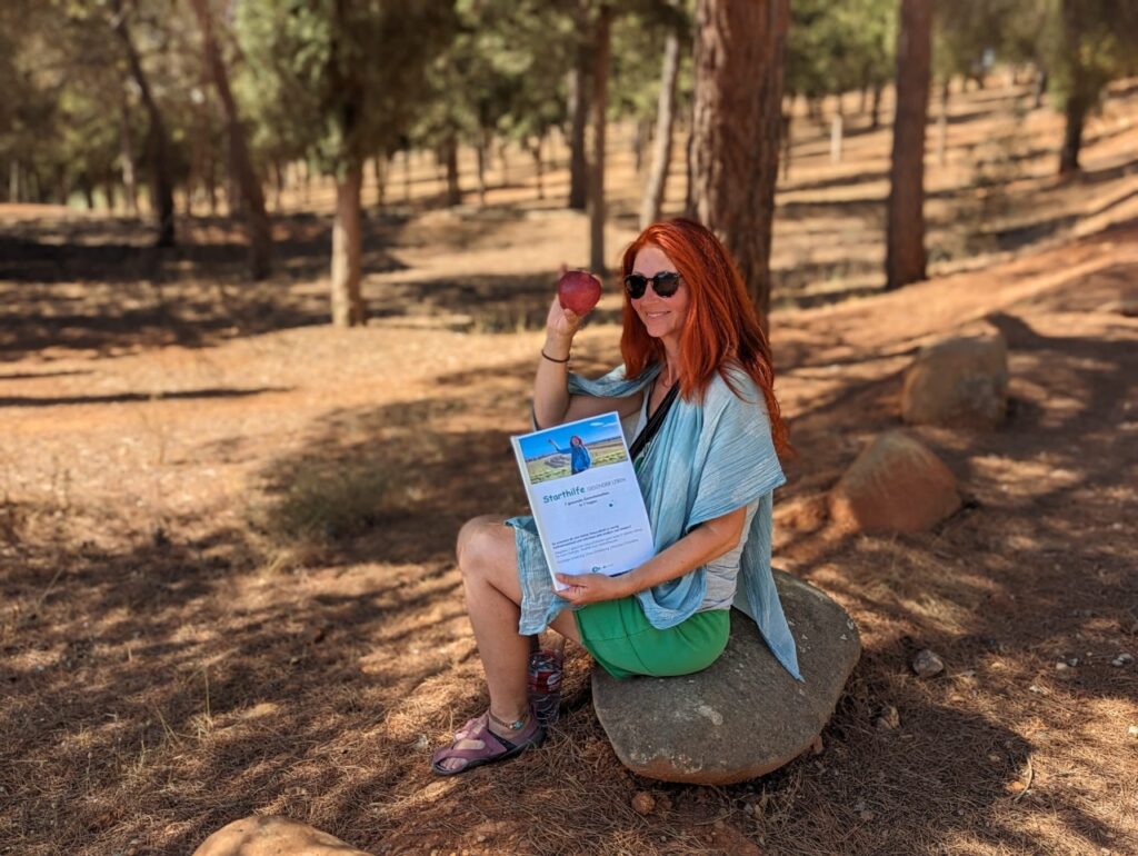 Karin sitzt mit Apfel und Starthilfe auf einem Stein im Wald mit roter Erde.