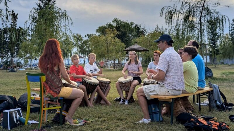 Eine Trommelgruppe sitzt im Kreis, zwischen den Knien Djemben, die am Boden stehen. Im HIntergrund sieht man die Liegewiese des Seebades