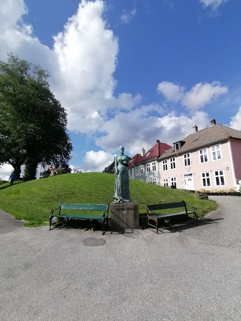 Grüne Hügelfläche, davor 2 Parkbänke, Links ein Baum, rechts ein Haus