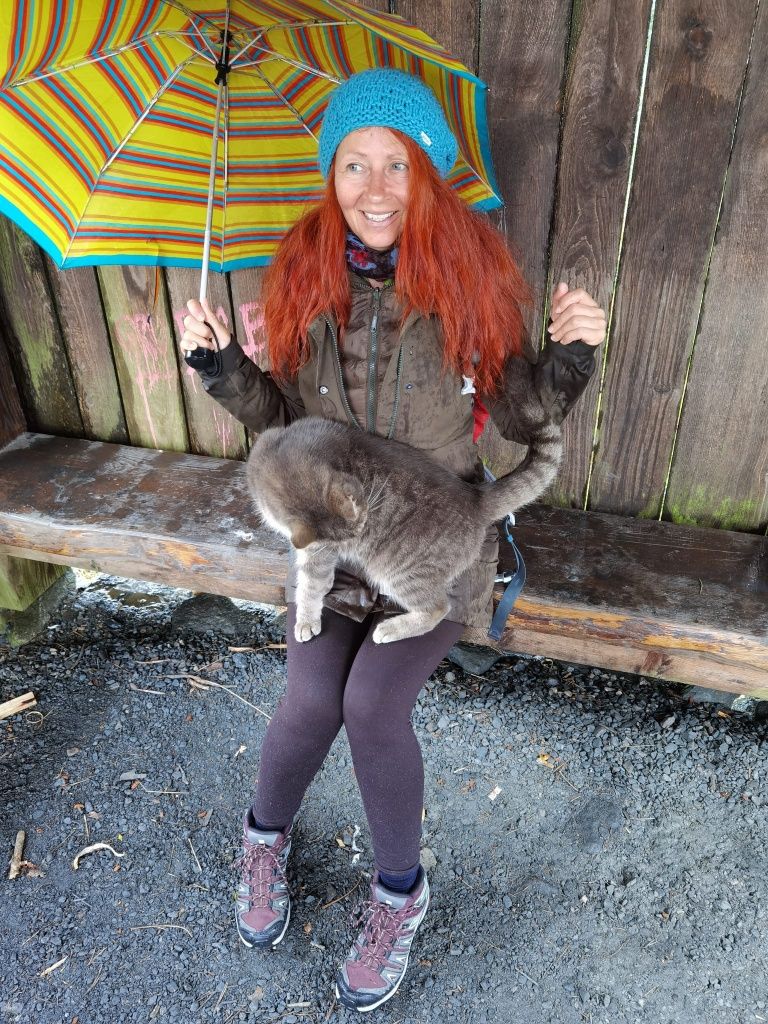 Karin mit Regenschirm, sitzend unter einem Holzunterstand, mit einer getigerten Katze am Schoss und einem skeptischen Gesichtsausdruck