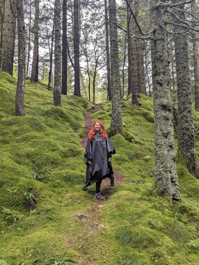 Karin mit schwarzem Regencape beim Aufstieg über den bemoosten Waldboden zwischen zahlreichen Bäumen