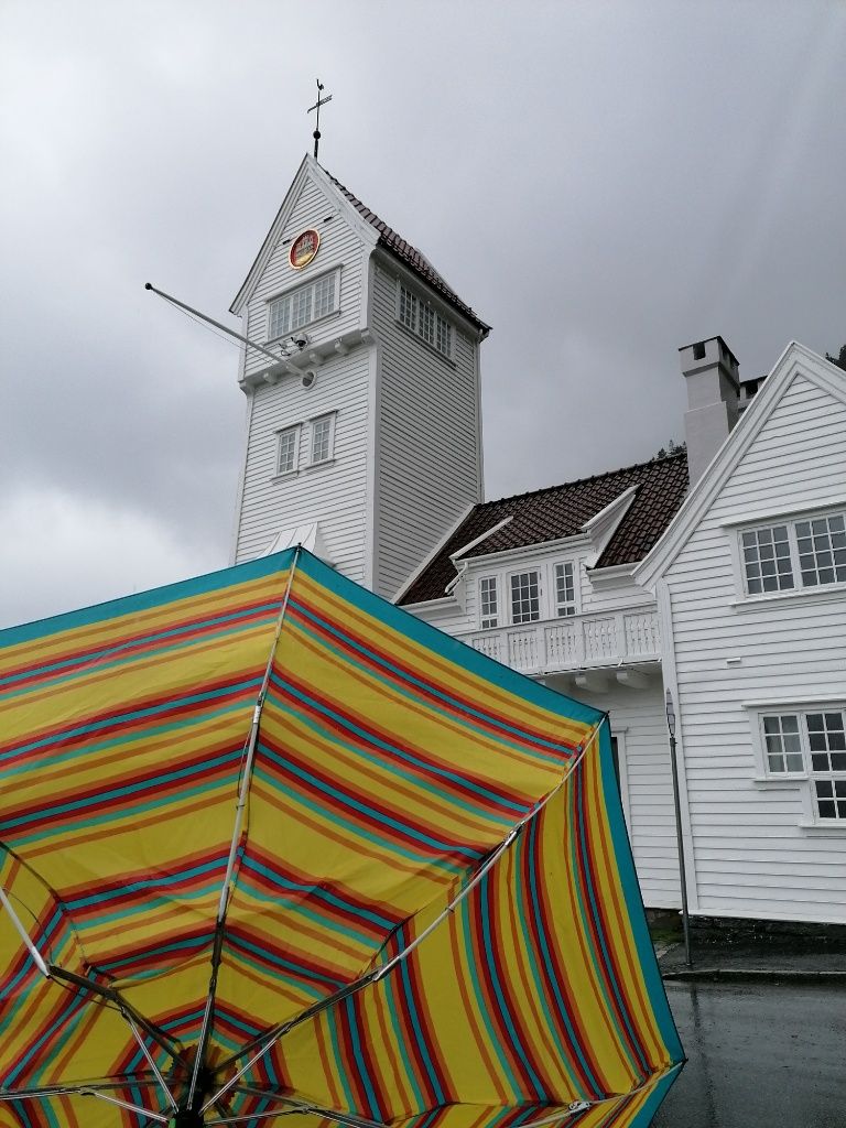 Vom Wind umgestülpter, bunt-gestreifter Regenschirm, im Hintergrund weißes Haus mit Turm